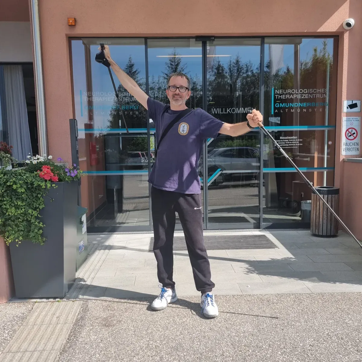Marc steht mit erhobenen Armen und zwei Stöcken vor dem Eingang der Rehaklinik, beim zweiten Aufenthalt ohne Rollstuhl